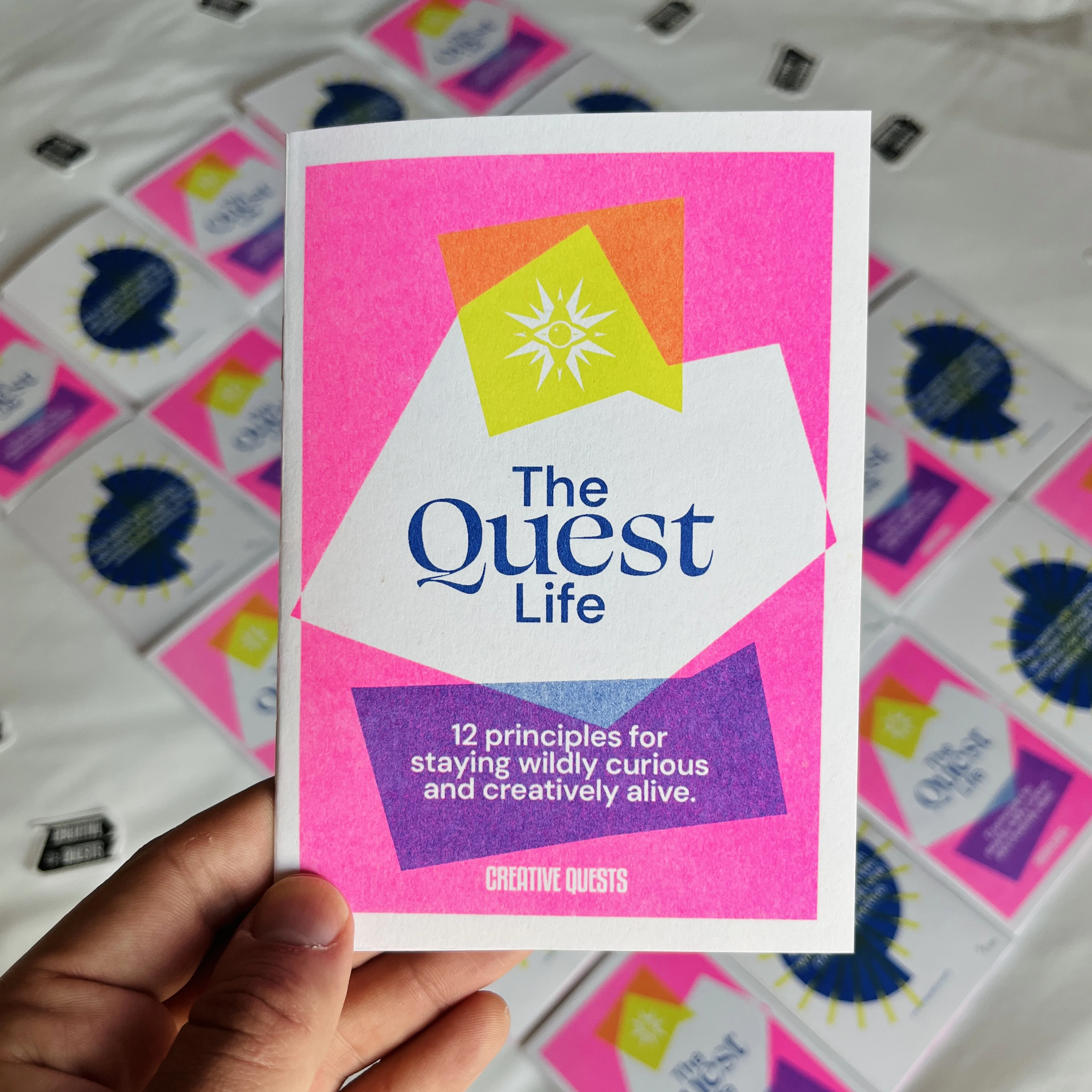 A hand holds a brightly coloured booklet titled “The Quest Life” with neon geometric shapes and the subtitle: “12 principles for staying wildly curious and creatively alive.” Copies of the booklet are scattered blurred in the background.