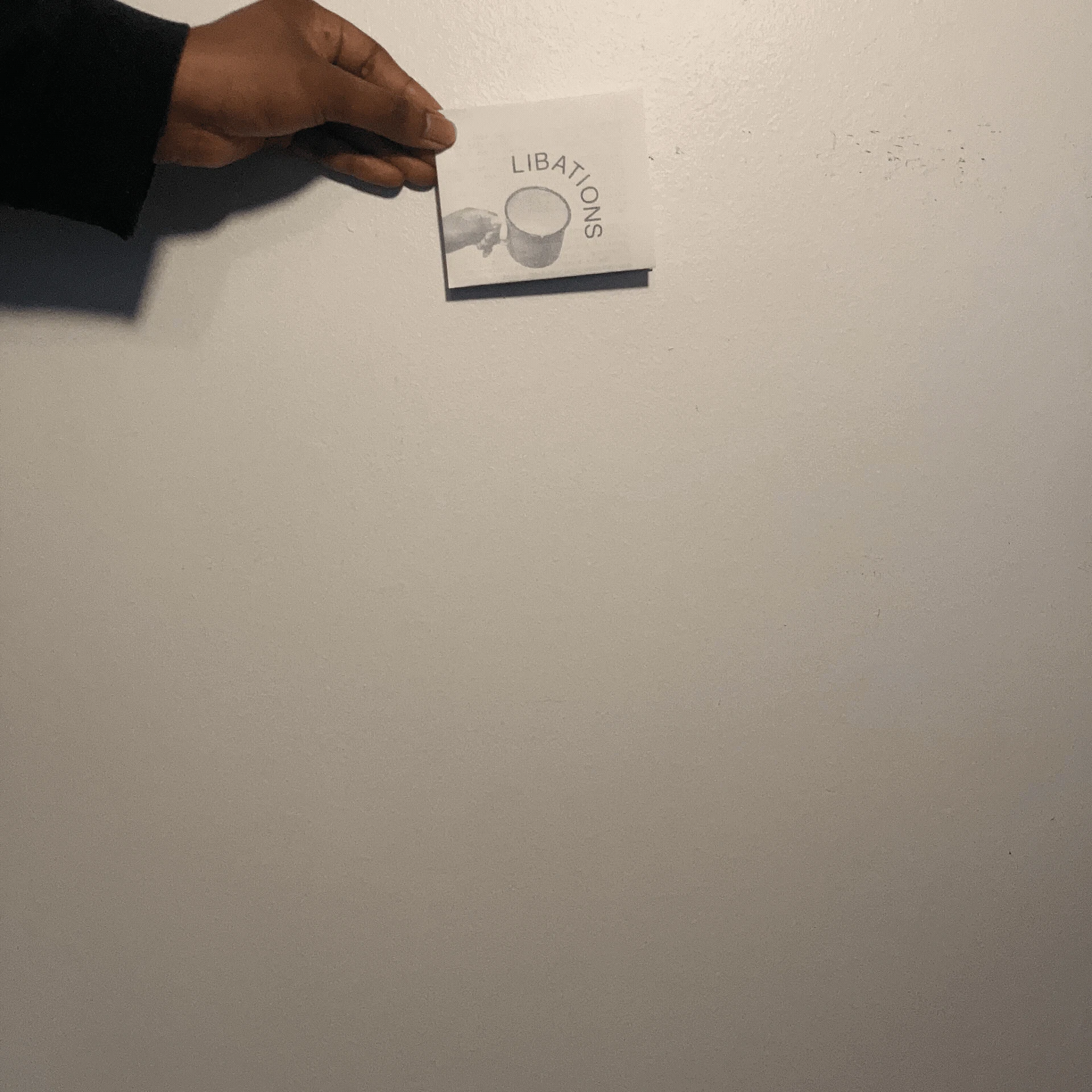 Beautiful brown hands hold and unfold an 11x17 inch zine. The cover shows the title "Libations" curving around an image of an enamel cup held by another beautiful hand.