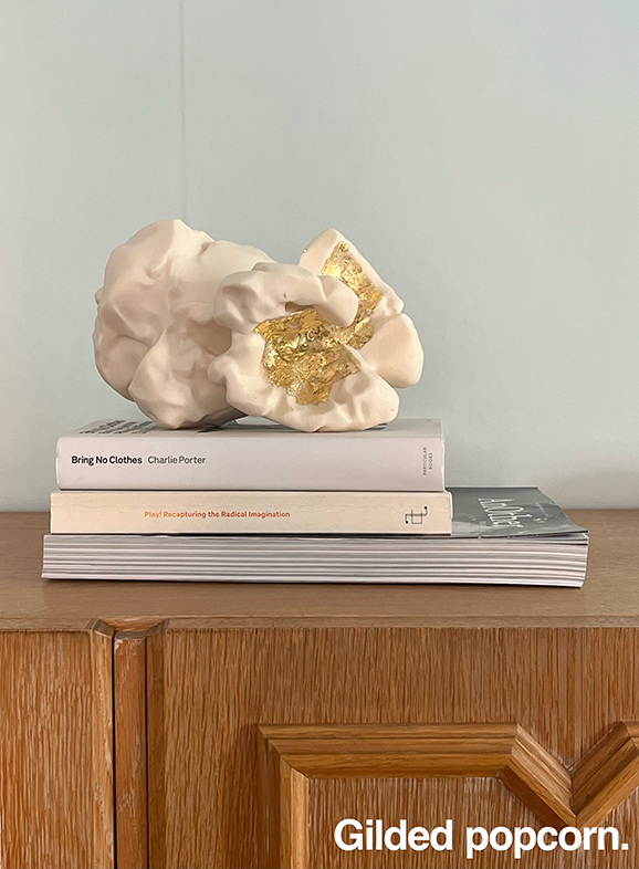 A larde ceramic popcorn with leaf gold kernel on top of three books resting on a shelf. The books are "Bring no clothes" by Clharlie Porter, "Play! Recapturing the Radical Imagination" and a magazine that appears to be named "AnOther".