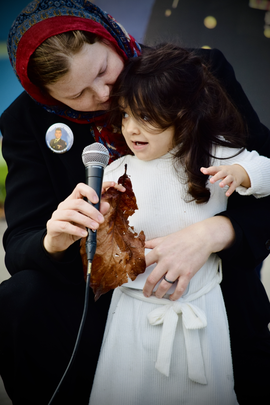 Melissa Chaudhry with young Salma holding a microphone, at the Armistice Day (Veterans Day) rally where she called for her husband's release. On Melissa's lapel is a button showing her husband's military portrait, which reads Free Zahid Now!