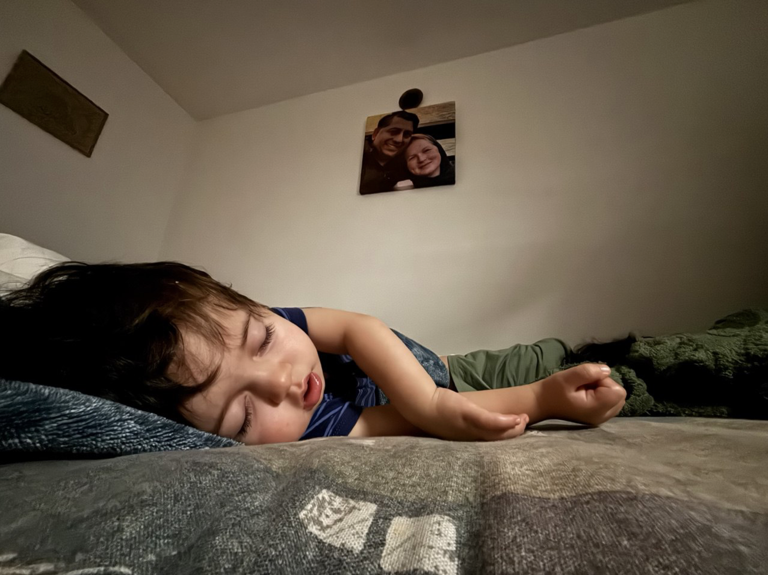 Adil asleep alone during his father's detention. A portrait of Zahid and Melissa in happier times hangs on the wall above him.