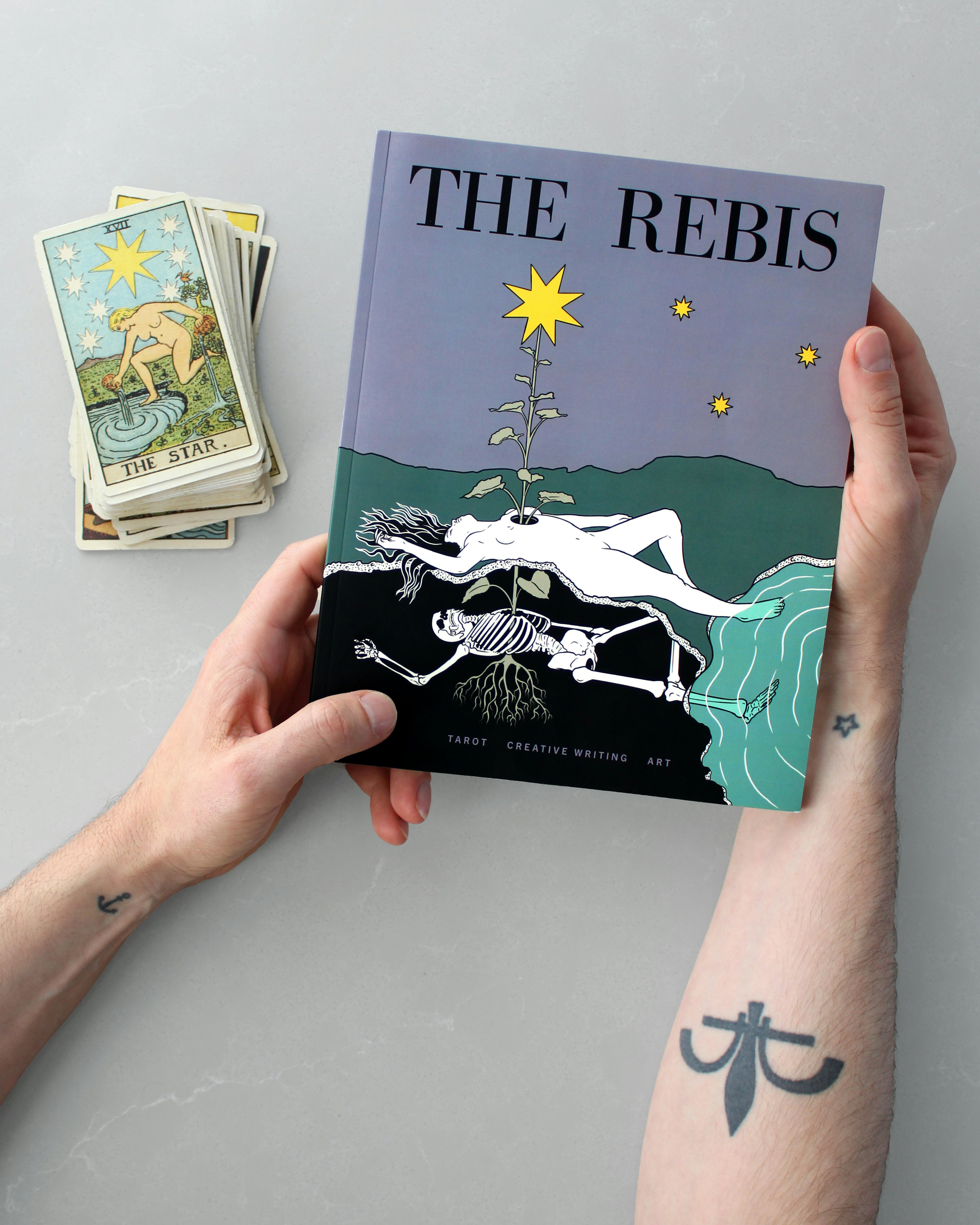 Two hands hold a print issue of The Rebis: The Star, next to a pack of tarot playing cards showing the Rider-Waite-Smith version of The Star archetype. On the cover of the magazine is a figure lying on the earth, with one foot in a pool of water. The figure's skeleton lies underneath, and a flower is growing through the skeleton and the figure.