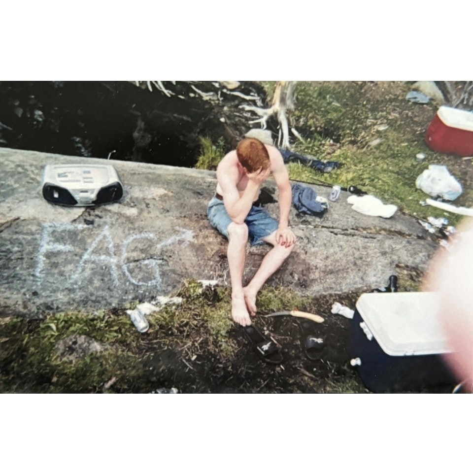This photo shows a shirtless young man with red hair sitting on a rock outdoors, covering his face with one hand as if upset or tired. He wears denim shorts and sits barefoot, sandals on the ground nearby. Around him are coolers, cans, a machete-like knife, a boombox, and scattered trash. Chalk writing on the rock spells a slur.