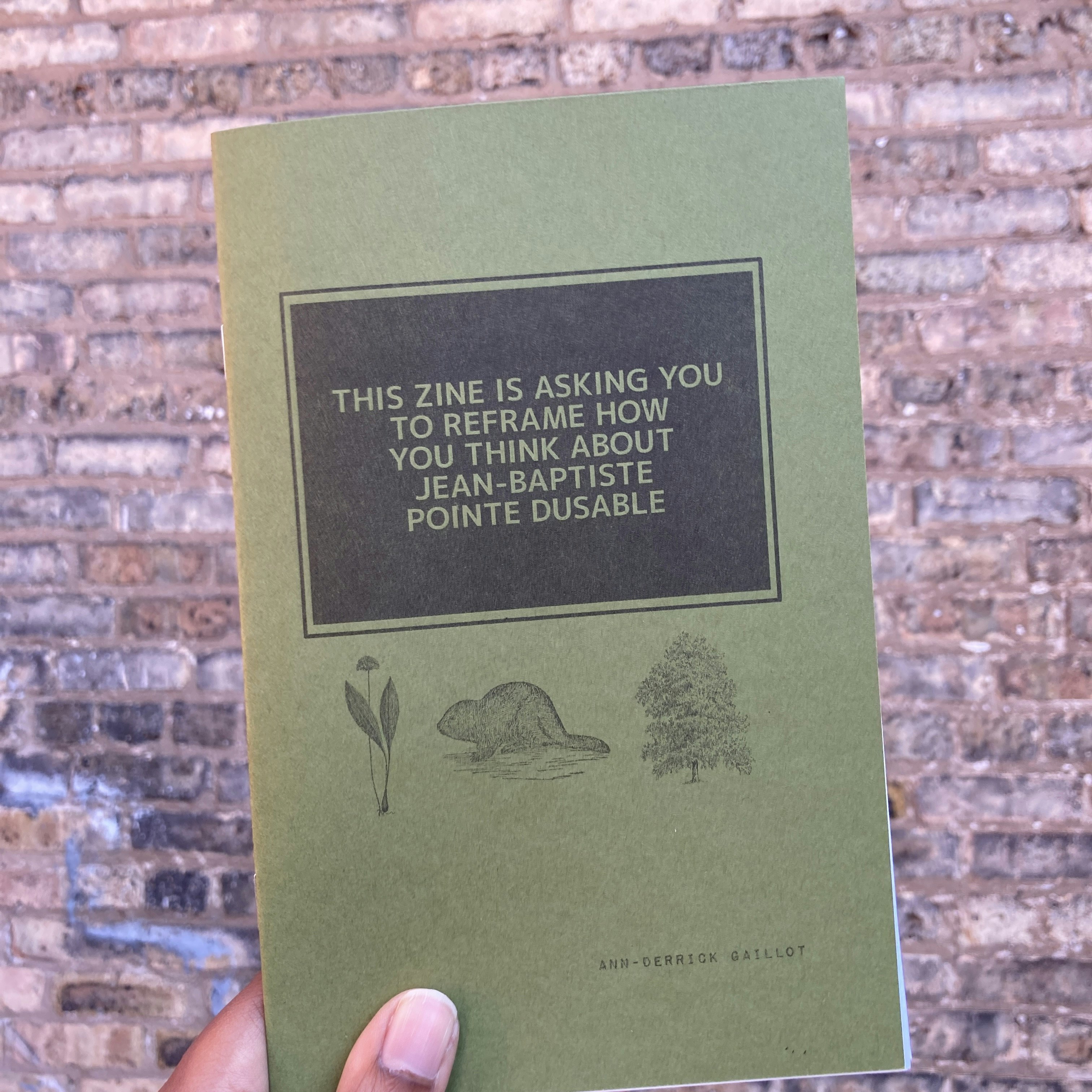 A hand holds a green pamphlet with the words 'THIS ZINE IS ASKING YOU TO REFRAME HOW YOU THINK ABOUT JEAN-BAPTISTE POINTE DUSABLE'. A brick wall is in the background.